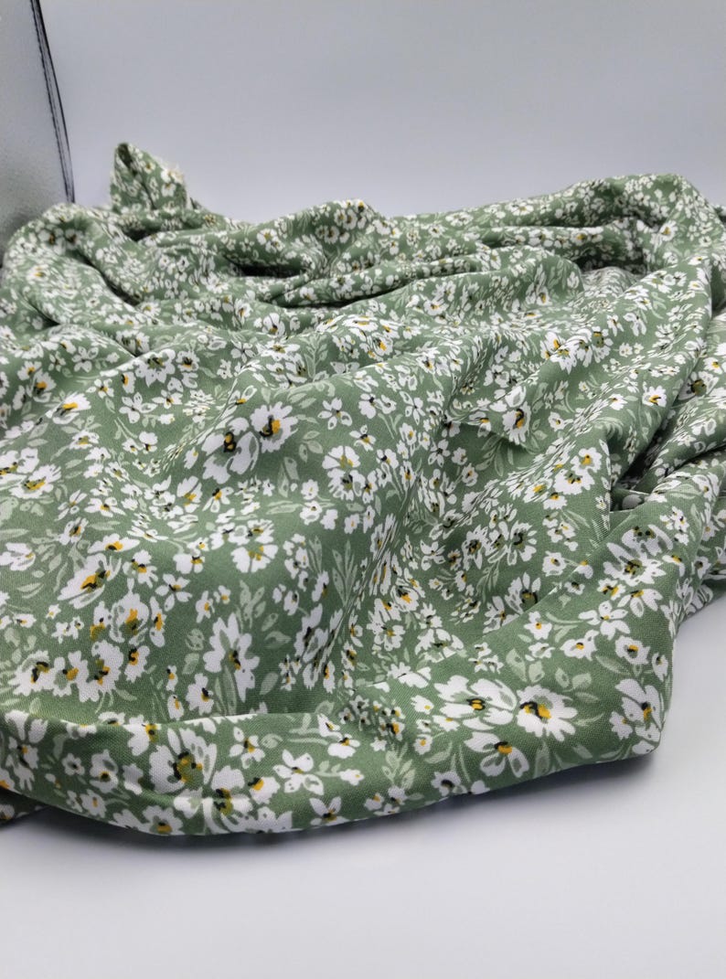 May include: A folded piece of fabric with a repeating floral pattern. The fabric is a muted green color, covered in small white flowers with yellow centers. The fabric appears soft and lightweight, suitable for clothing or home decor.