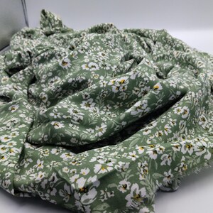 May include: A folded piece of fabric with a floral pattern. The fabric is a muted green color, covered in small white flowers with yellow centers. The fabric appears to be soft and lightweight, with a slightly frayed edge.