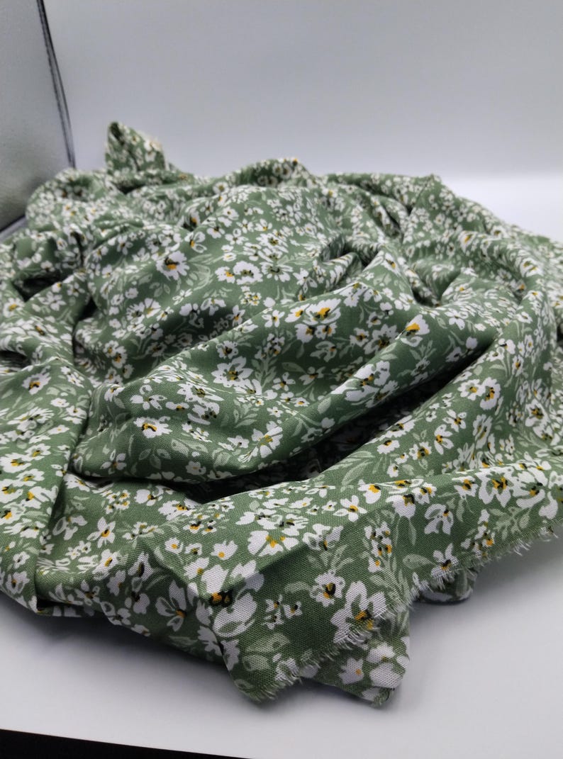 May include: A folded piece of fabric with a floral pattern. The fabric is a muted green color, covered in small white flowers with yellow centers. The fabric appears to be soft and lightweight, with a slightly frayed edge.