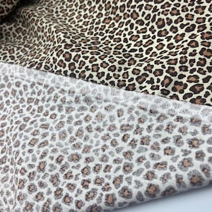 Puede incluir: Una pieza de tela doblada con un estampado de leopardo. La sección superior muestra un estampado animal negro y marrón de alto contraste sobre un fondo crema, mientras que la sección inferior muestra una versión más clara en gris y beige.