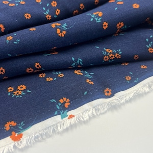 May include: A navy blue fabric featuring a repeating pattern of small orange flowers with teal green leaves. The material is folded in layers, revealing a white, frayed selvage edge along the bottom corner of the textile piece.