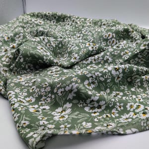 May include: Close-up of a green fabric with a white floral pattern. The fabric is draped, showing the texture and design. The flowers have yellow centers, and the leaves are a darker green.