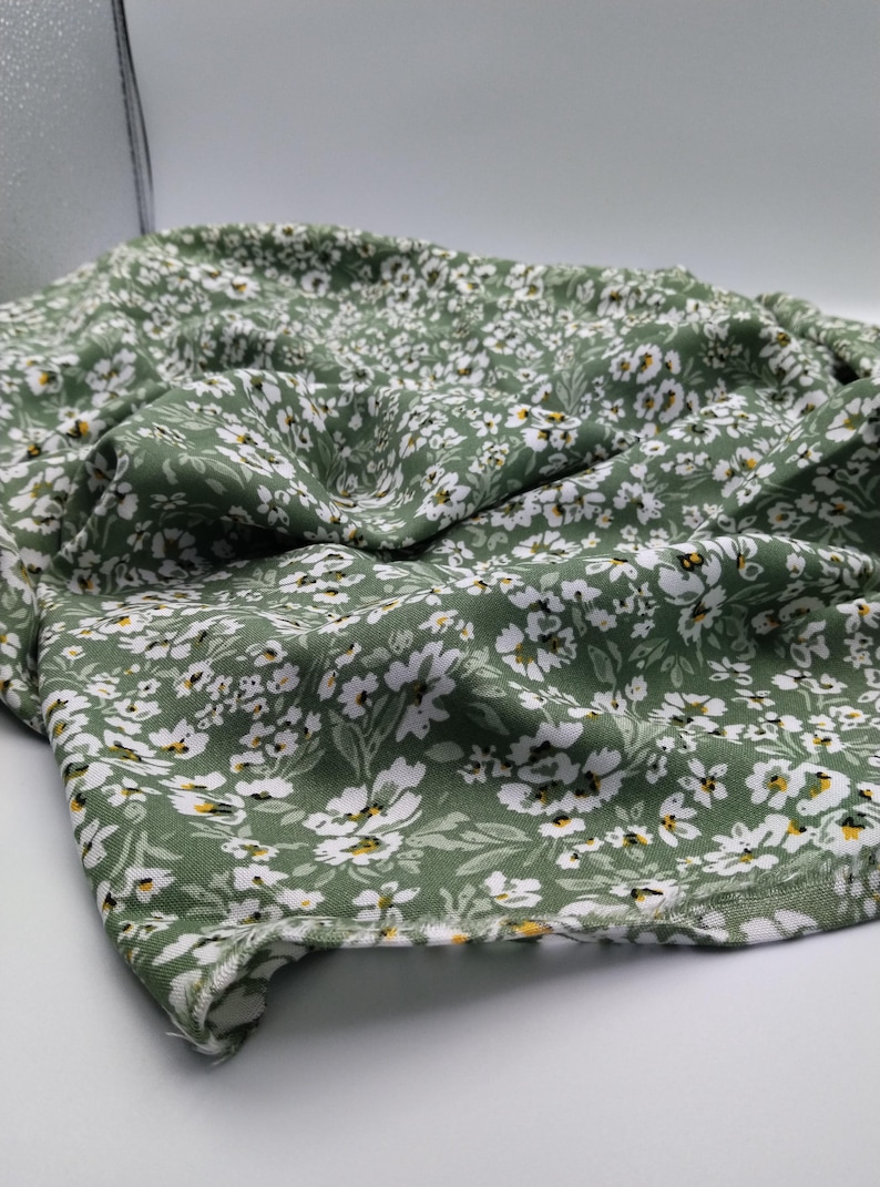 May include: Close-up of a green fabric with a white floral pattern. The fabric is draped, showing the texture and design. The flowers have yellow centers, and the leaves are a darker green.