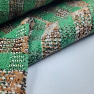 May include: A woven fabric featuring a plaid pattern in vibrant green, brown, and white. The material has a textured, fuzzy surface with visible interlaced threads and a loose, frayed edge showing the construction of the weave and fibers.