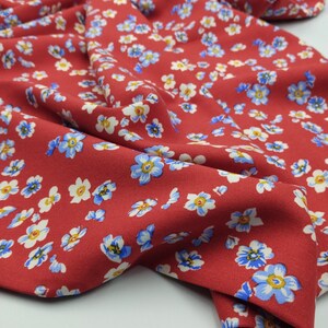 K&ouml;nnte beinhalten: Nahaufnahme eines roten Stoffes mit Blumenmuster. Der Stoff ist mit kleinen blauen, wei&szlig;en und gelben Blumen bedeckt. Der Stoff ist gefaltet und zeigt die Textur und die Details des Blumendrucks.