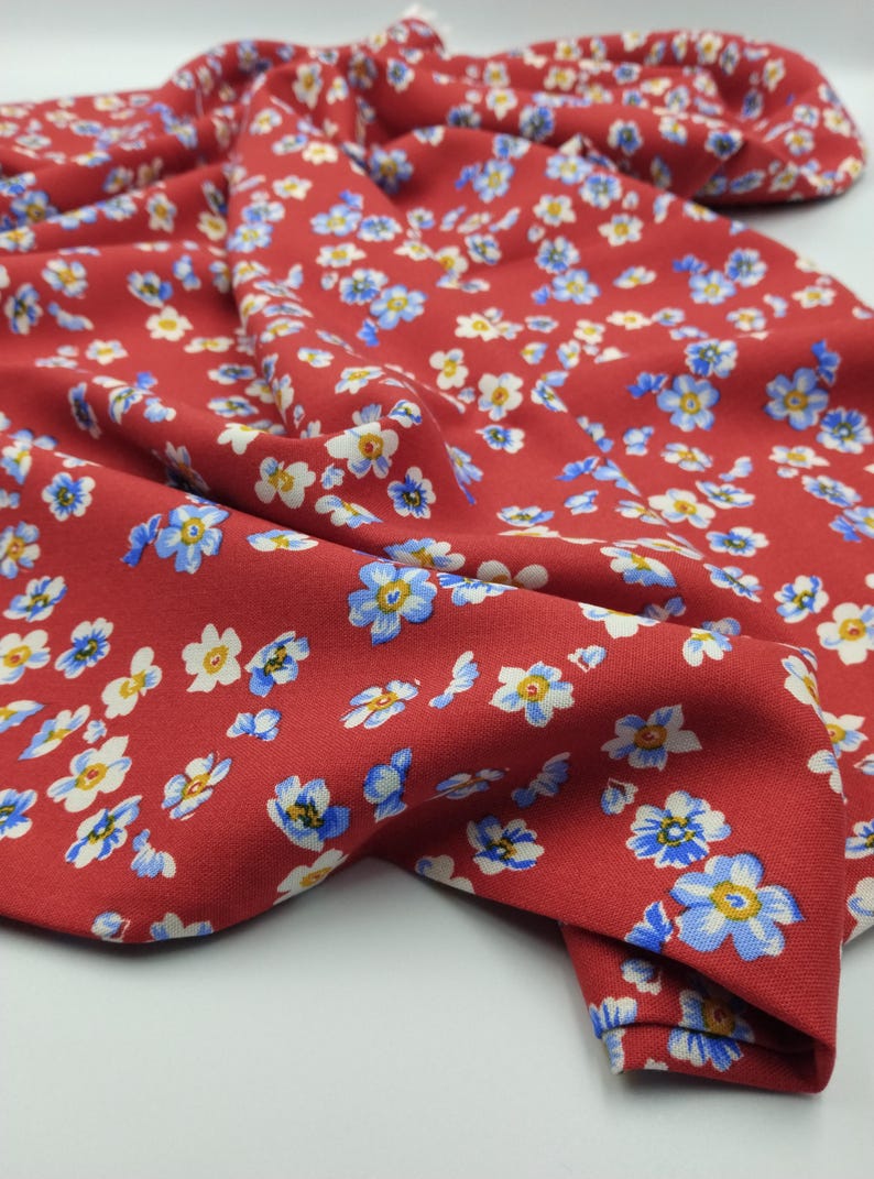 K&ouml;nnte beinhalten: Nahaufnahme eines roten Stoffes mit Blumenmuster. Der Stoff ist mit kleinen blauen, wei&szlig;en und gelben Blumen bedeckt. Der Stoff ist gefaltet und zeigt die Textur und die Details des Blumendrucks.
