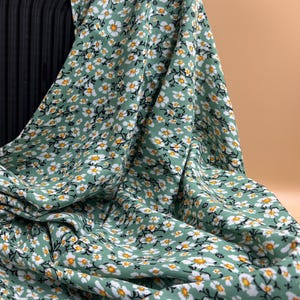 May include: A sage green fabric featuring a repeating pattern of small white flowers with yellow centers and thin black stems. The material is draped in soft folds, showcasing the dense floral print across the entire surface of the textile.