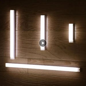 Op de afbeelding: Vier witte LED-lichtstrips gemonteerd op een houten muur. De strips zijn van verschillende lengtes, waarbij de langste strip horizontaal aan de onderkant van de afbeelding is gemonteerd.