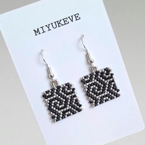 May include: A pair of square beaded earrings with a black and silver pattern. The earrings are hanging from silver hooks. The text "MIYUKEVE" is printed on the white card behind the earrings.