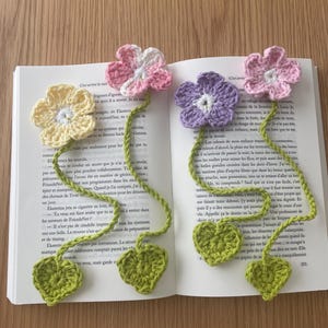 Peut inclure: Quatre marque-pages faits à la main en crochet en forme de fleurs jaunes, roses, violettes et roses. Chaque marque-page a une tige verte et une base en forme de feuille verte. Les marque-pages sont placés sur un livre ouvert.