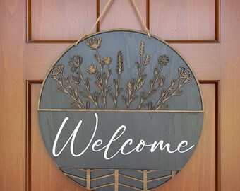 May include: Round wooden welcome sign with a gray background and floral design. The word "Welcome" is written in white script. The sign is hanging on a wooden door.