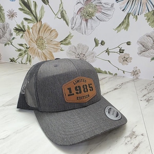 May include: A gray trucker hat with a black mesh back. The front of the hat features a brown leather patch with the text "LIMITED 1985 EDITION". A small sticker is on the brim.