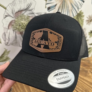 May include: Black snapback hat with a brown leather patch featuring the letter "A" and the word "AGUAYO". The hat has a mesh back and a curved brim. The hat has a "Snapback" tag.