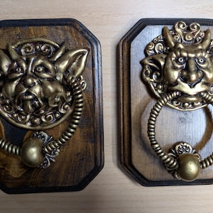 May include: Two decorative door knockers. Each knocker features a brass-colored goblin face with a ring handle. The knockers are mounted on stained wooden plaques with a dark border. The goblin faces have intricate details.