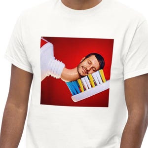 Op de afbeelding: Een wit t-shirt met een vierkante grafische print op de borst. De afbeelding toont een hoofd dat rust op een grote tandenborstelkop met blauwe, gele en witte borstelharen, tegen een effen, felrode achtergrond.
