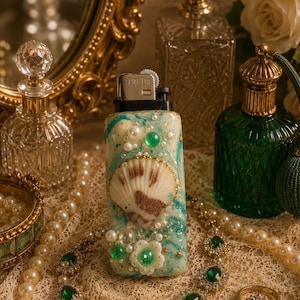 May include: A rectangular lighter featuring a marbled teal and white surface, decorated with a central seashell, small white pearls, green beads, and tiny gold star accents. The top metal component is engraved with the text SPIN-LITE.