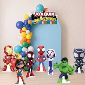 May include: A party decoration set featuring a blue arch backdrop with a balloon garland in blue, yellow, orange, green, and white. Includes cardboard cutouts of Iron Man, Violet Parr, Miles Morales, Spider-Man, Ghost-Spider, Hulk, and Black Panther.