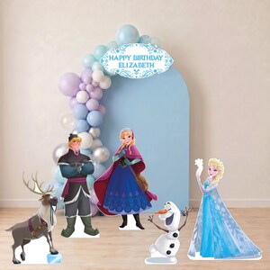 May include: A party decoration set featuring cardboard cutouts of Frozen characters including Kristoff, Anna, Olaf, and Elsa, alongside a blue arched backdrop with a balloon garland and a sign that reads HAPPY BIRTHDAY ELIZABETH in blue text.