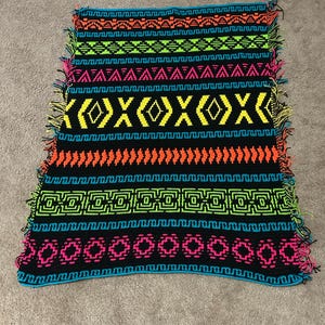 May include: A rectangular crochet blanket featuring a black base with horizontal bands of neon orange, lime green, hot pink, and bright blue geometric patterns. The edges are finished with multi-colored fringe made of yarn strands.