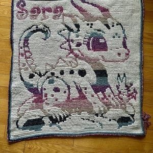 May include: A rectangular crochet blanket featuring a white background with a large, stylized dragon graphic in shades of purple, blue, and black. The name "Sora" is crocheted in purple letters in the upper left corner. The edges have a dark border.