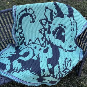 May include: A crocheted blanket in shades of teal and gray, featuring a pixelated design of a sea creature. The blanket is draped over a dark gray chair, with a detailed border in coordinating colors. The blanket is handmade.