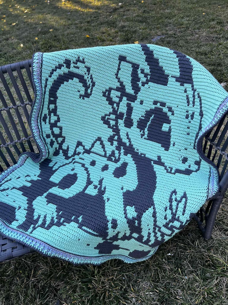 May include: A crocheted blanket in shades of teal and gray, featuring a pixelated design of a sea creature. The blanket is draped over a dark gray chair, with a detailed border in coordinating colors. The blanket is handmade.
