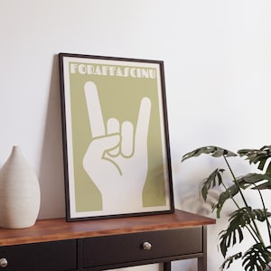 Può includere: Un poster incorniciato con uno sfondo verde chiaro e un gesto della mano bianco, il simbolo 'rock on'. Il testo "FORAFFA SCINU" è in alto. Il poster è in una cornice nera e poggia su un tavolo di legno.