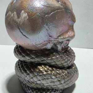 May include: A sculpture of a human skull atop a coiled snake. The skull has a metallic, iridescent finish with shades of brown, pink, and silver. The snake is a dark, textured gray with a scale pattern.