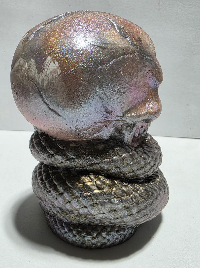 May include: A sculpture of a human skull atop a coiled snake. The skull has a metallic, iridescent finish with shades of brown, pink, and silver. The snake is a dark, textured gray with a scale pattern.
