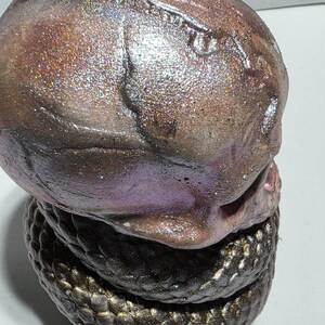 May include: A sculpted, abstract art piece featuring a textured, iridescent head atop a coiled base. The head transitions from brown to purple with glittery accents. The base is a dark, scaled pattern.