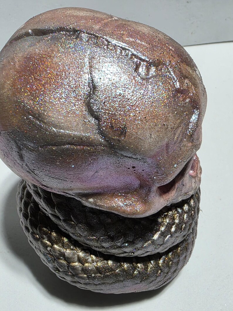 May include: A sculpted, abstract art piece featuring a textured, iridescent head atop a coiled base. The head transitions from brown to purple with glittery accents. The base is a dark, scaled pattern.