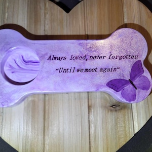 May include: A purple bone-shaped memorial plaque with the words "Always loved, never forgotten" and "Until we meet again." A purple butterfly is painted on the right side. The plaque is set on a wooden heart-shaped base.