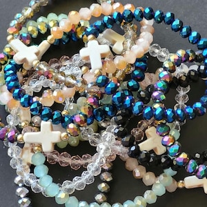 May include: A collection of elastic bracelets featuring faceted glass beads in various colors including blue, black, peach, clear, and light green. Each bracelet is accented with a white stone cross bead, creating a layered and textured appearance.