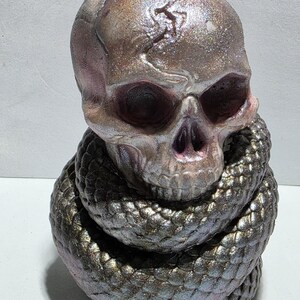 May include: A metallic skull sculpture with a snake coiled around its base. The skull has a crack on top and is painted in shades of pink, purple, and silver. The snake is a dark, iridescent color, with scales that catch the light.