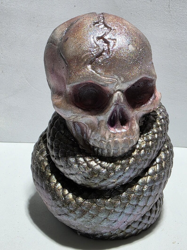 May include: A metallic skull sculpture with a snake coiled around its base. The skull has a crack on top and is painted in shades of pink, purple, and silver. The snake is a dark, iridescent color, with scales that catch the light.