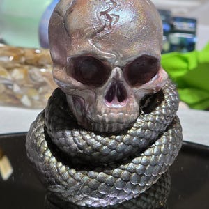 May include: A detailed sculpture of a human skull with a snake coiled around its base. The skull has a cracked surface and is painted with iridescent colors. The snake is dark gray with scale details.