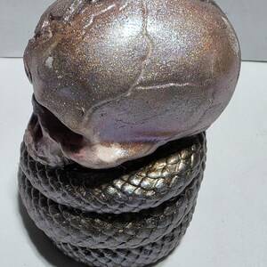 May include: A ceramic sculpture of a skull atop a coiled snake. The skull has a metallic, iridescent finish with shades of brown, purple, and silver. The snake is dark with a textured, scale-like pattern.