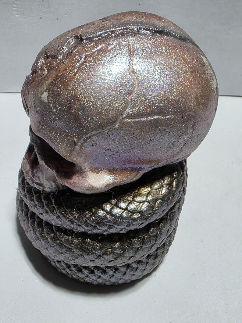 May include: A ceramic sculpture of a skull atop a coiled snake. The skull has a metallic, iridescent finish with shades of brown, purple, and silver. The snake is dark with a textured, scale-like pattern.