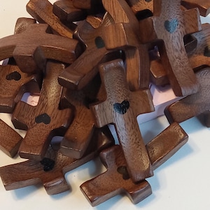 May include: A pile of small, wooden crosses with a dark brown finish. Each cross has a small, black heart painted on it. The crosses are stacked together, creating a textured surface.