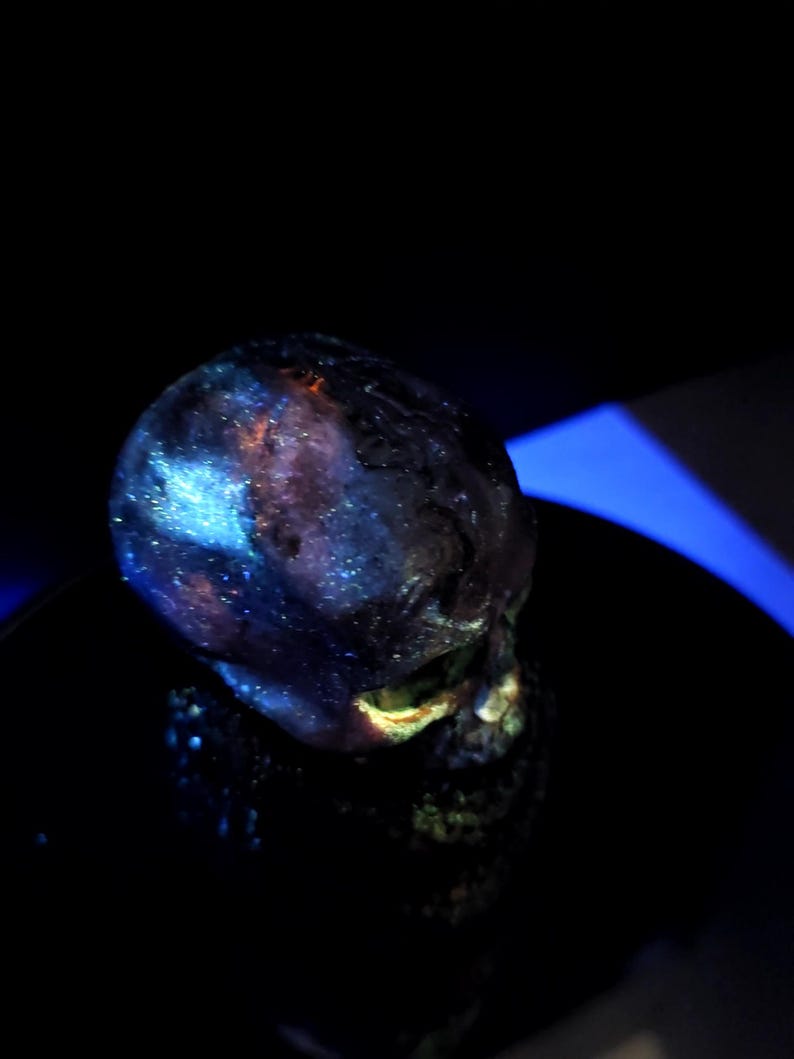 May include: A dark, glowing skull sculpture with a galaxy-like pattern of blues, purples, and oranges. The skull is set against a black background, with a hint of blue light.