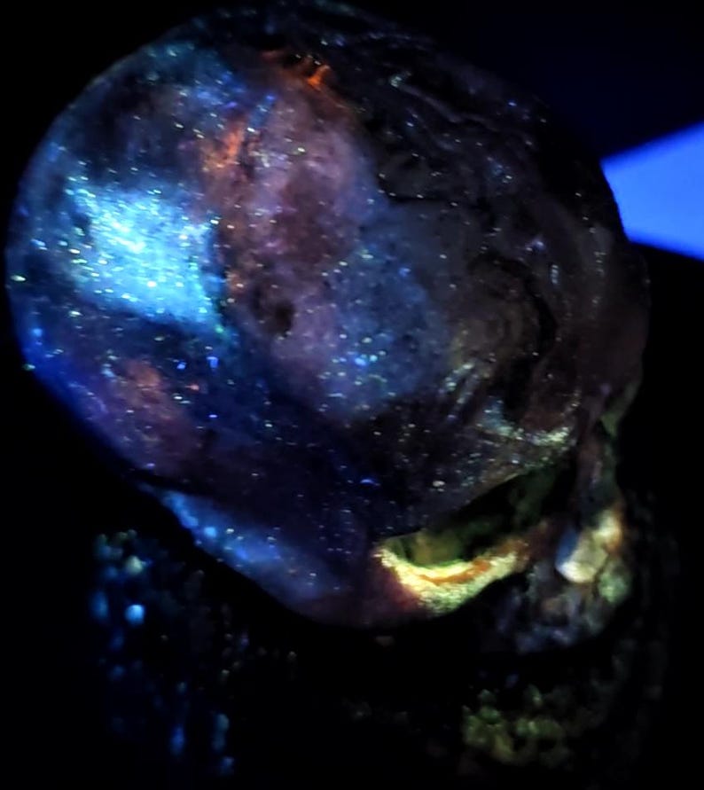 May include: A glowing, iridescent skull with a dark background. The skull has a blue and purple color scheme with flecks of white and orange. The jawline is highlighted with yellow and orange.