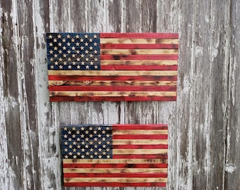 May include: Two wooden American flags are shown, one large and one small. The flags have the traditional design of red and white stripes and a blue section with white stars. The wood has a distressed, rustic appearance.