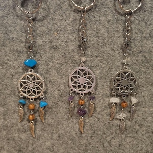 Puede incluir: Cinco llaveros de atrapasueños plateados con cuentas y plumas de diferentes colores. Los atrapasueños tienen un círculo plateado con un diseño de estrella en el centro. Las cuentas son rosas, azules, moradas, blancas y negras.