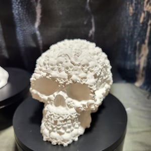 May include: A white skull sculpture with a textured surface, featuring numerous smaller skulls covering the head. The sculpture is displayed on a black rotating stand, showcasing its intricate details and design.