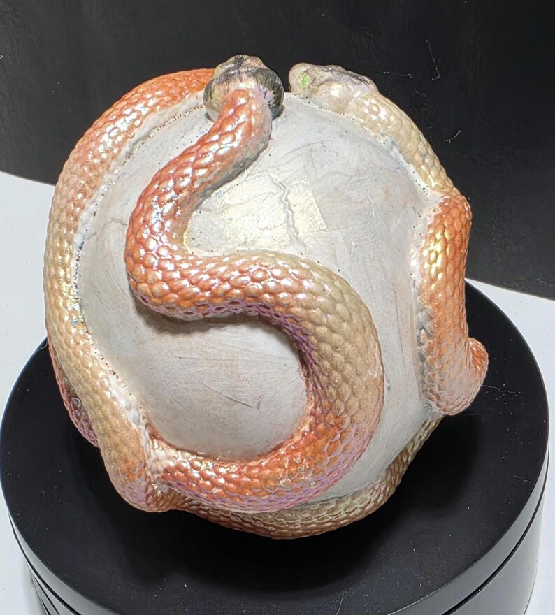Puede incluir: Una escultura de una esfera blanca con dos serpientes enrolladas alrededor. Las serpientes tienen escamas iridiscentes en tonos naranja, rosa y dorado. La escultura est&aacute; sobre una base negra.