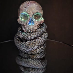 May include: A detailed sculpture of a skull with iridescent green and blue accents. The skull is wrapped in a coiled, textured snake. The sculpture is set against a dark background, creating a dramatic effect.