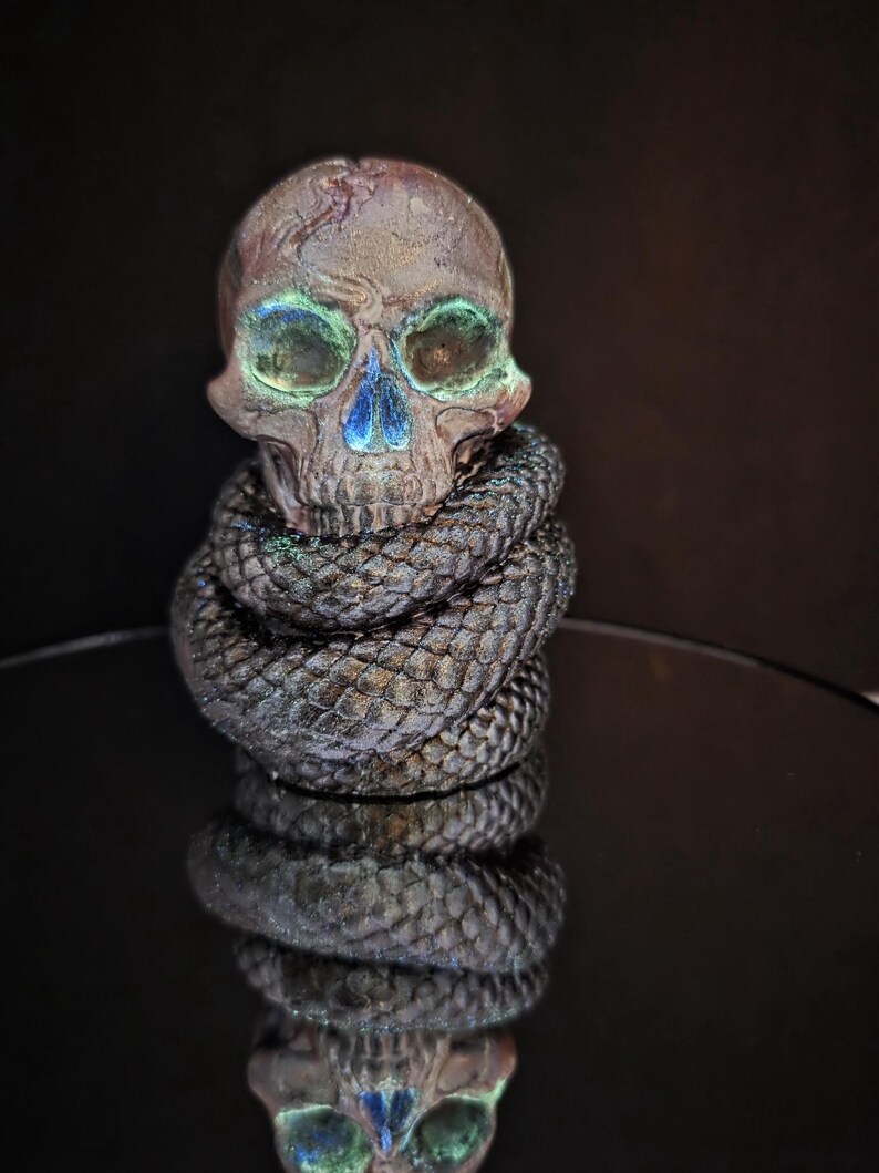 May include: A detailed sculpture of a skull with iridescent green and blue accents. The skull is wrapped in a coiled, textured snake. The sculpture is set against a dark background, creating a dramatic effect.