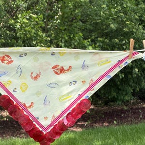 Może przedstawiać: Trójkątna bandana z jasnego żółtego materiału z powtarzającym się nadrukiem czerwonych homarów, krewetek i różnych muszli. Krawędzie wykończone są czerwono-białą wstążką w kratkę oraz rzędem półprzezroczystych, czerwonych, okrągłych cekinów.