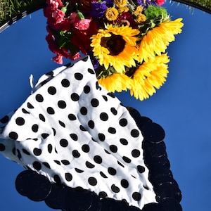 May include: A white fabric accessory featuring a black polka dot pattern and a border of circular black sequins. The item is positioned on a reflective surface alongside a bouquet of yellow sunflowers, red carnations, and purple filler flowers.