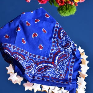 Puede incluir: Un pañuelo azul brillante con un estampado de cachemira en blanco y rojo. Los bordes de la tela están adornados con una serie de pequeños dijes en forma de estrella de color claro, hechos de un material nacarado liso, creando un borde decorativo.